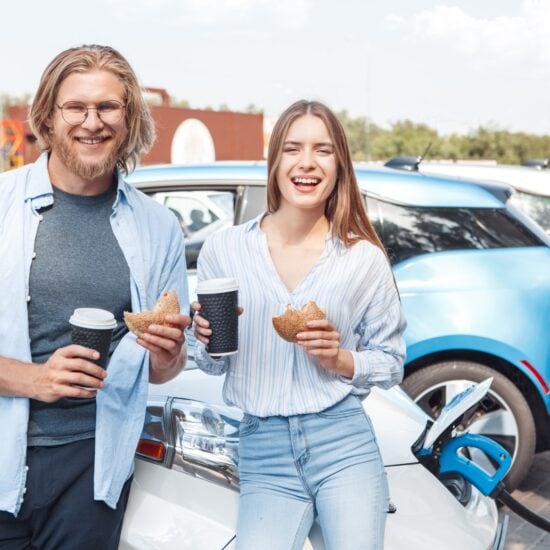 Twee mensen staan met een kopje koffie en een bagel in hun hand bij een wegrestaurant in Duitsland. Achter hen staat een elektrische auto op te laden.