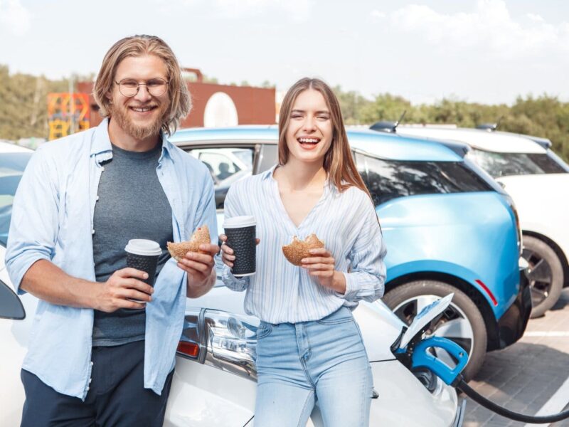 Twee mensen staan met een kopje koffie en een bagel in hun hand bij een wegrestaurant in Duitsland. Achter hen staat een elektrische auto op te laden.