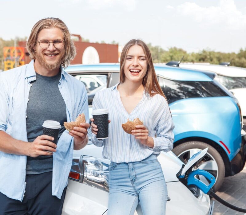 Twee mensen staan met een kopje koffie en een bagel in hun hand bij een wegrestaurant in Duitsland. Achter hen staat een elektrische auto op te laden.