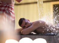 Een man in een rood-wit geruite handdoek giet water over een andere man heen. Deze behandeling wordt watertherapie genoemd en wordt uitgevoerd in een traditionele Turkse hamam.