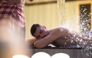 Een man in een rood-wit geruite handdoek giet water over een andere man heen. Deze behandeling wordt watertherapie genoemd en wordt uitgevoerd in een traditionele Turkse hamam.
