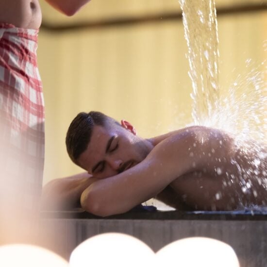 Een man in een rood-wit geruite handdoek giet water over een andere man heen. Deze behandeling wordt watertherapie genoemd en wordt uitgevoerd in een traditionele Turkse hamam.