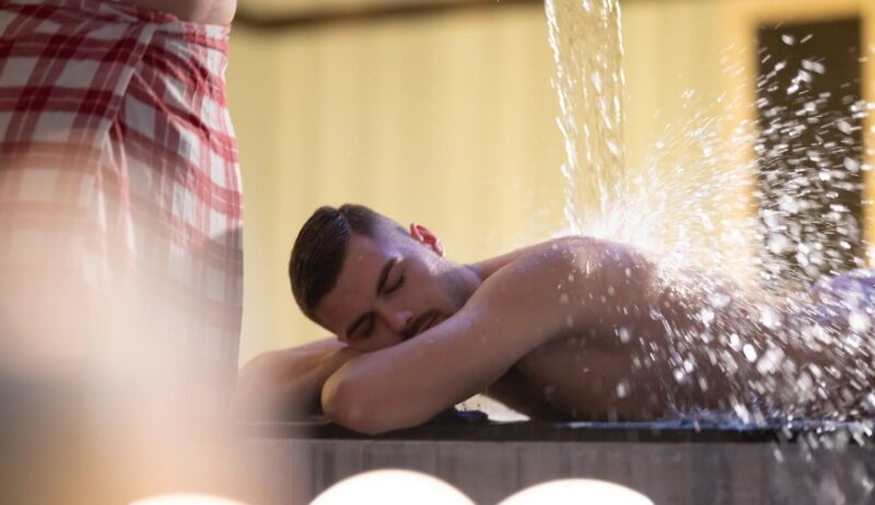 Een man in een rood-wit geruite handdoek giet water over een andere man heen. Deze behandeling wordt watertherapie genoemd en wordt uitgevoerd in een traditionele Turkse hamam.