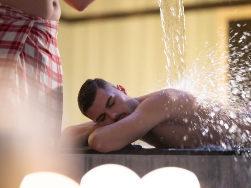 Een man in een rood-wit geruite handdoek giet water over een andere man heen. Deze behandeling wordt watertherapie genoemd en wordt uitgevoerd in een traditionele Turkse hamam.