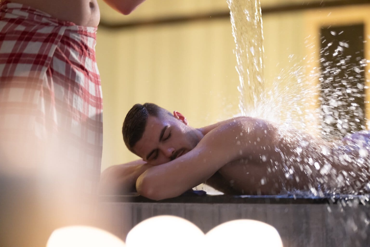 Een man in een rood-wit geruite handdoek giet water over een andere man heen. Deze behandeling wordt watertherapie genoemd en wordt uitgevoerd in een traditionele Turkse hamam.