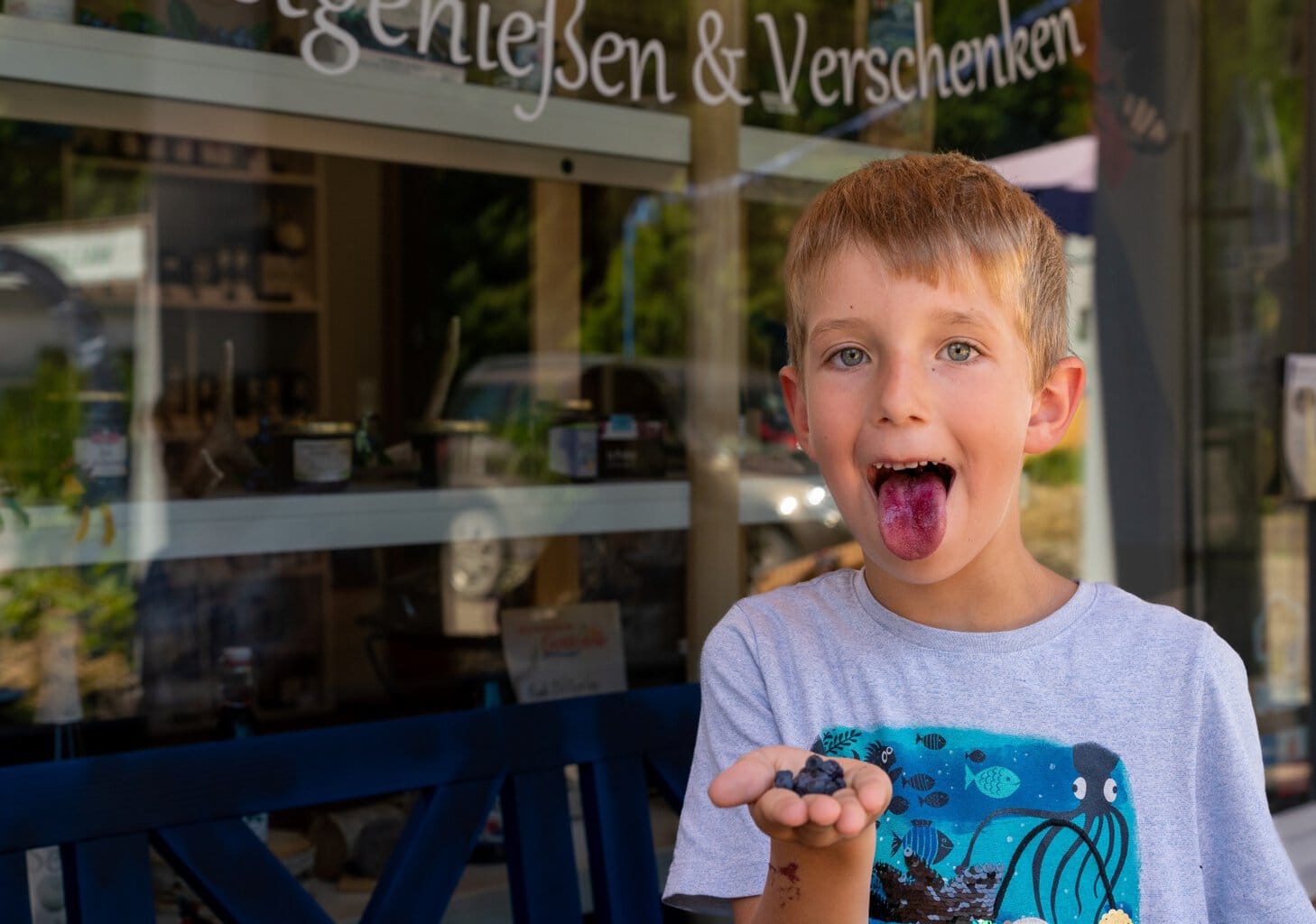 Lars, een jongen, steekt zijn paarse tong uit. In zijn hand houdt hij een tros bosbessen vast