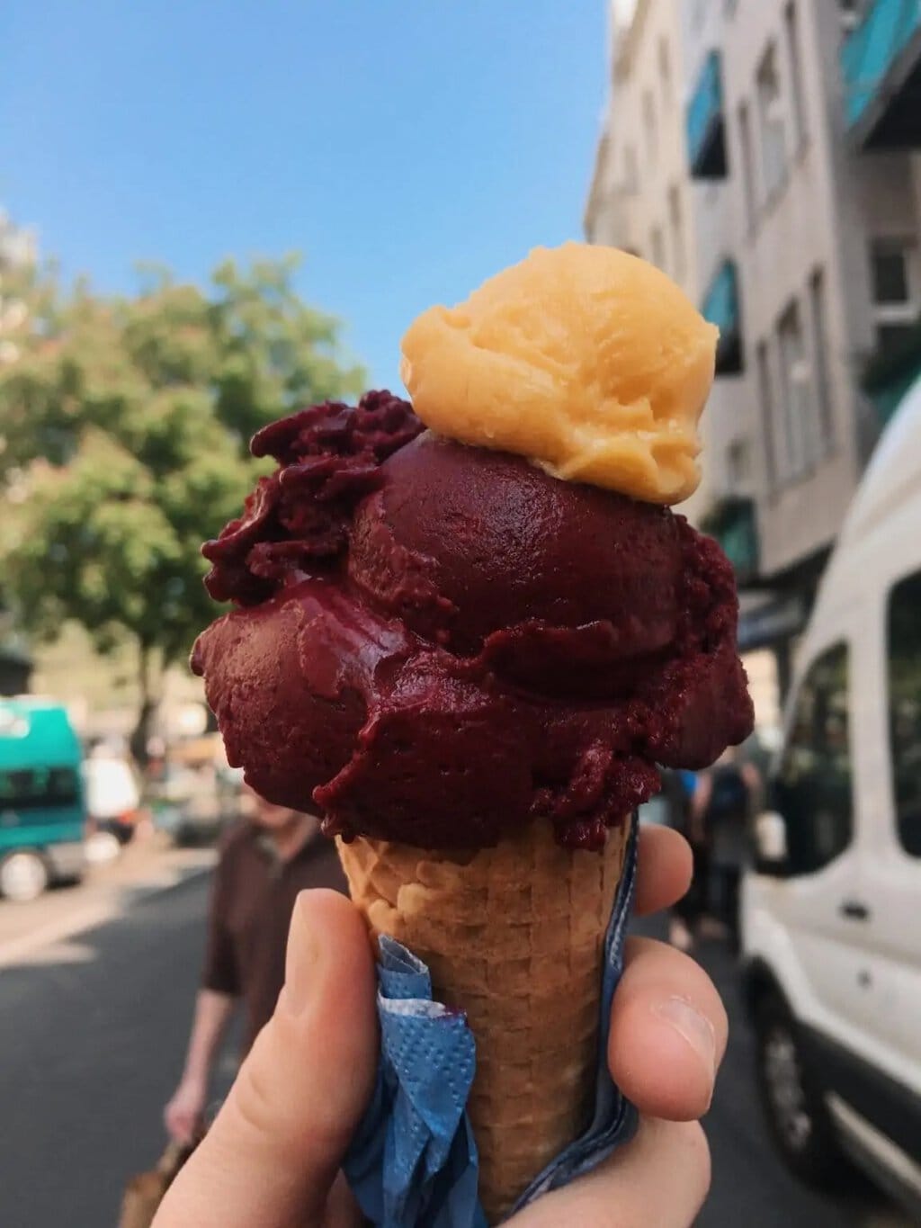Twee bolletjes ijs in een hoorntje met een blauw servetje. Op de achtergrond een straat in Keulen