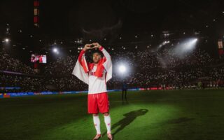 Foto uit de Netflix-documentaire ‘Poldi’: voetballer Lukas Podolski staat in het stadion op het veld in het wit-rode shirt van 1. FC Köln en vormt met zijn vingers een hartje.