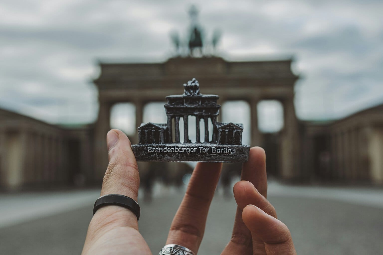 Een hand met een zwarte ring om de duim houdt een souvenir van de Brandenburger Tor voor de echte Brandenburger Tor
