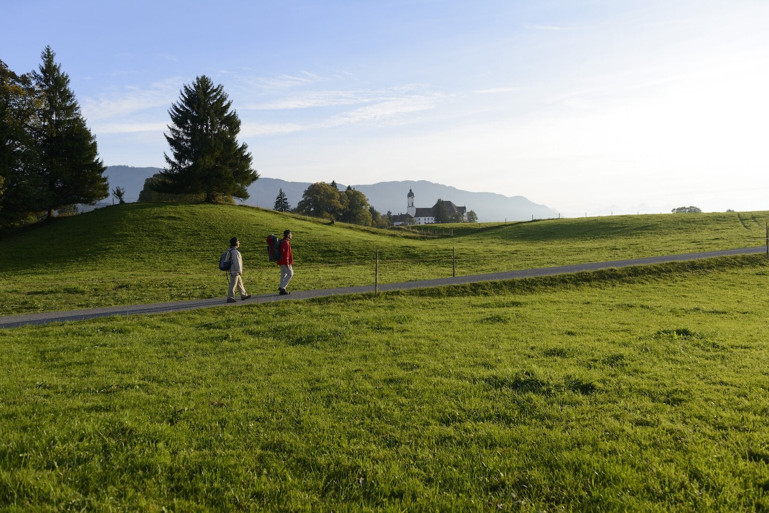 Twee wandelaars lopen over een pad tussen groene weiden. Op de achtergrond is de Wieskirche te zien 