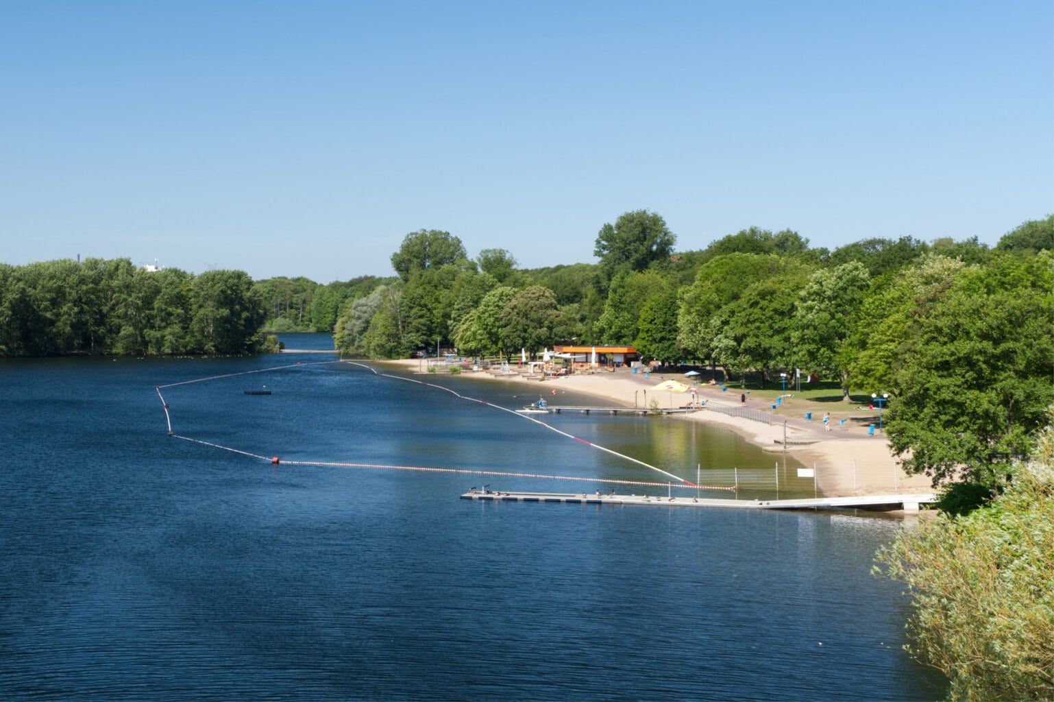 Een lang zandstrand bij een bosmeer, waar meerdere mensen liggen te zonnen.