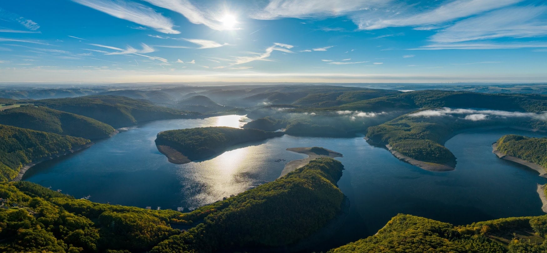 Een groot meer waarin meerdere landtongen uitsteken, vanuit de lucht gezien. Het landschap rond het meer is begroeid met dichte bossen en heuvelachtig, op het meer en in de dalen hangt nog wat mist. De hemel daarboven is stralend blauw, de zon wordt weerspiegeld door het wateroppervlak.