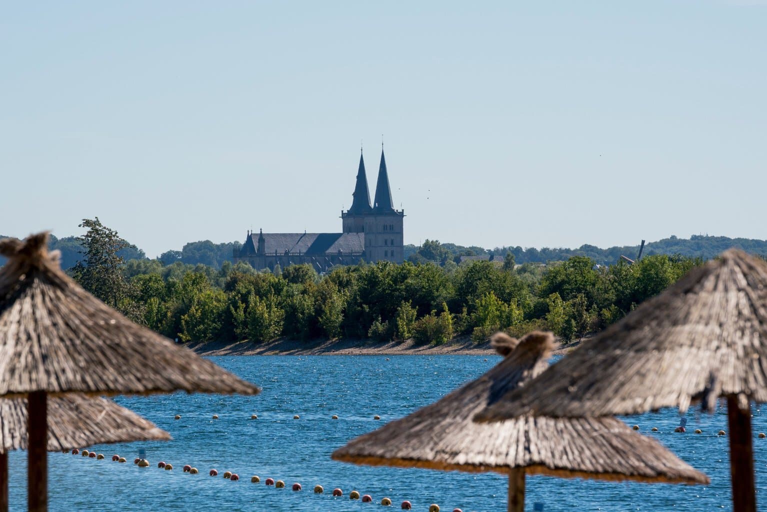 Boven een paar rieten strandschermen zweeft de blik over de Xantener Südsee naar de Dom van Xanten.