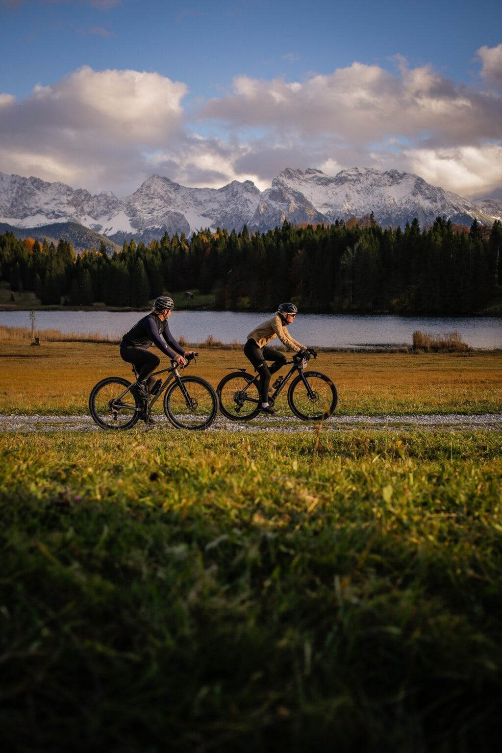 Twee personen rijden op gravelbikes op een onverharde weg langs een meer, met op de achtergrond een besneeuwde bergketen in de Alpen.
