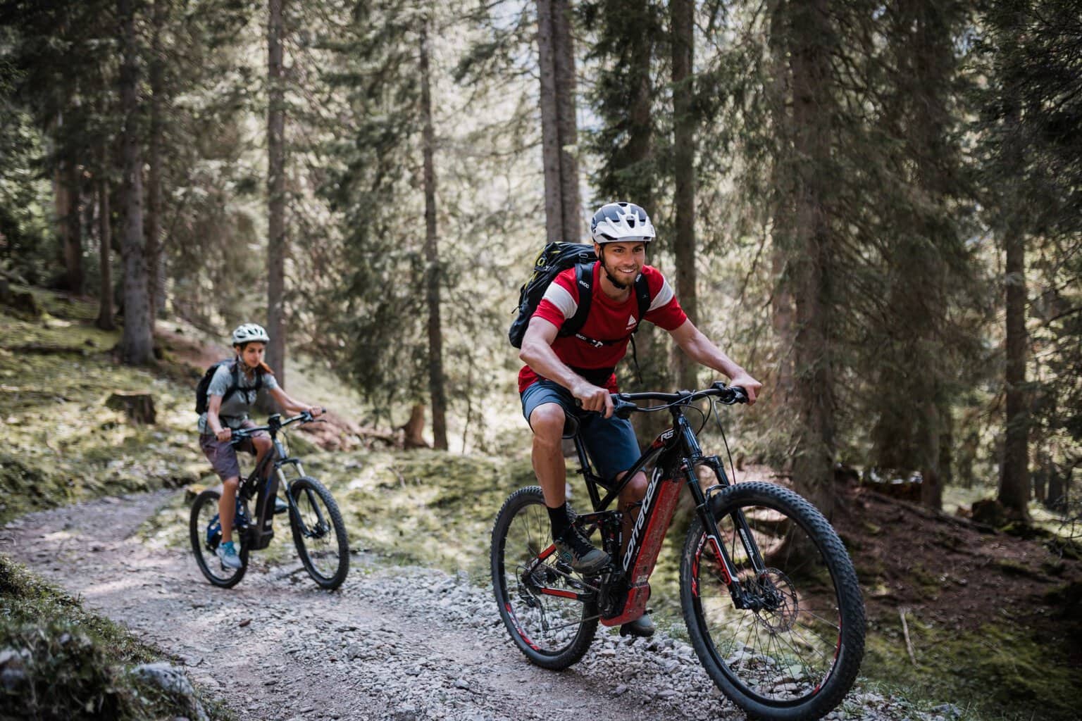 Een jonge man en een kind rijden met elektrische mountainbikes over een piste in een naaldbomenbos omhoog.