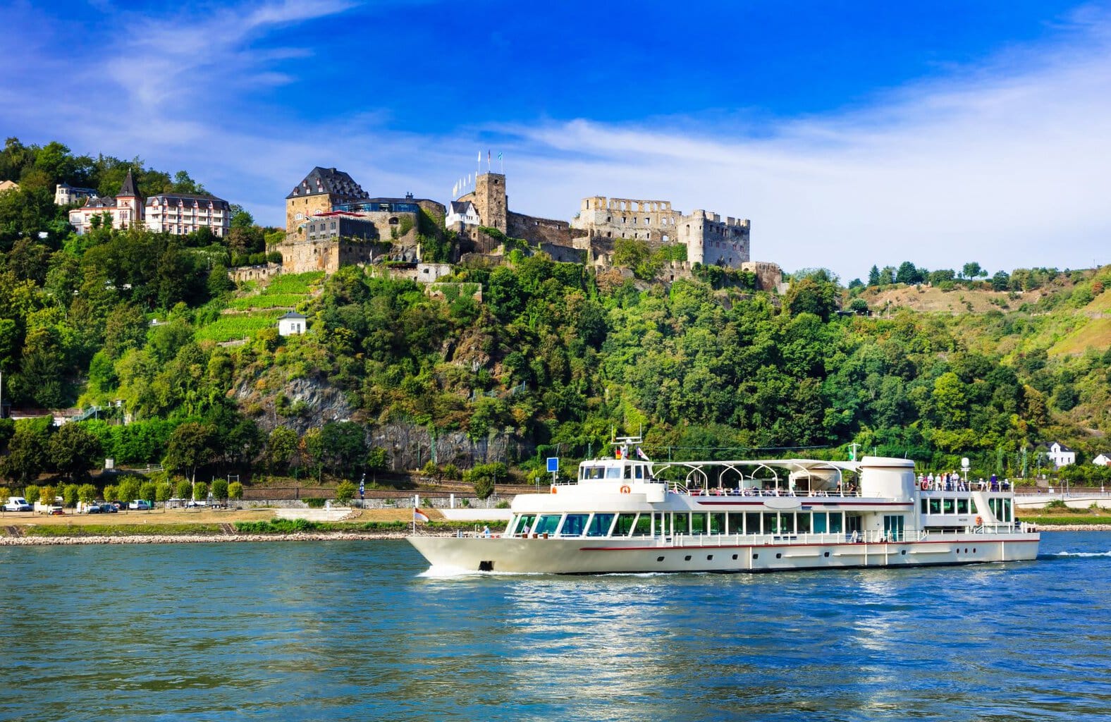Op de Rijn bij St. Goar is een witte veerboot zichtbaar: op de tegenoverliggende oever steekt de ruïne van de burcht Rheinfels met het aanpalende Schlosshotel Rheinfels uit een klein stuk bos.
