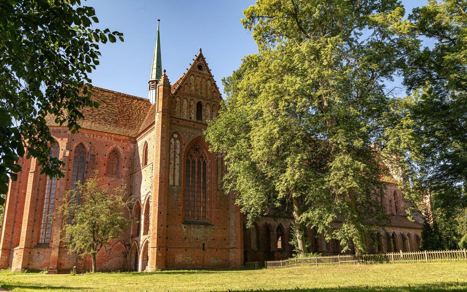 Het gotische bakstenen klooster Chorin in Brandenburg met zijn sierlijke gevel en slanke daktorentje, omringd door bomen en weiden onder een blauwe hemel.