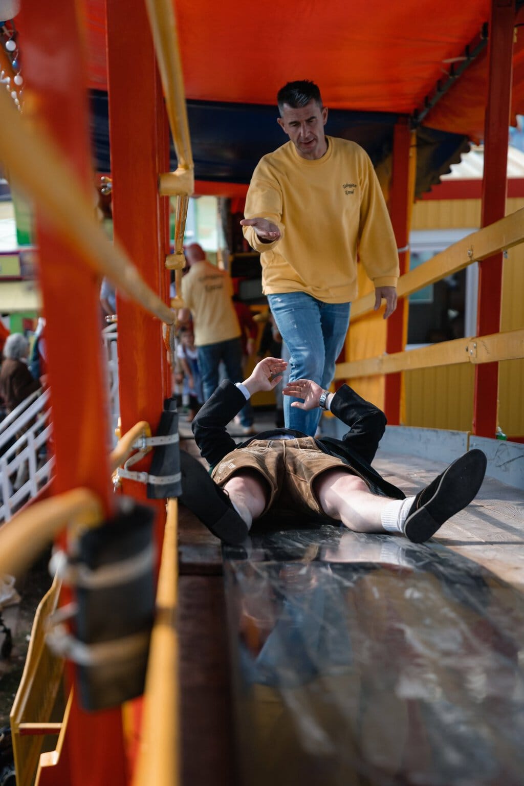 Man in lederhosen ligt onderaan de klassieke glijbaan Toboggan op het Oktoberfest, een medewerker helpt hem overeind