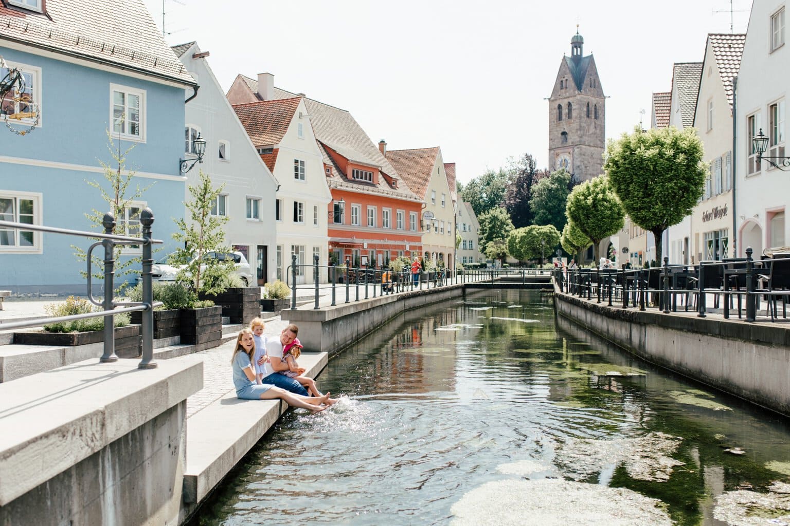 Gezin met twee kleine kinderen laat de voeten bungelen in de stadsbeek van Memmingen, met de toren van de Frauenkirche op de achtergrond, een ontspannen uitje met kinderen in Beieren
