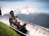 Vader en dochter lachend op de zomerrodelbaan bij Berchtesgaden met de besneeuwde Watzmann op de achtergrond, een spannend uitje met kinderen in Beieren