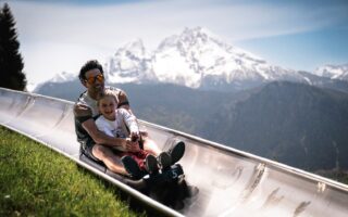 Vader en dochter lachend op de zomerrodelbaan bij Berchtesgaden met de besneeuwde Watzmann op de achtergrond, een spannend uitje met kinderen in Beieren