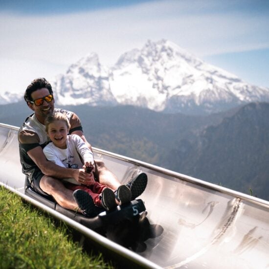 Vader en dochter lachend op de zomerrodelbaan bij Berchtesgaden met de besneeuwde Watzmann op de achtergrond, een spannend uitje met kinderen in Beieren
