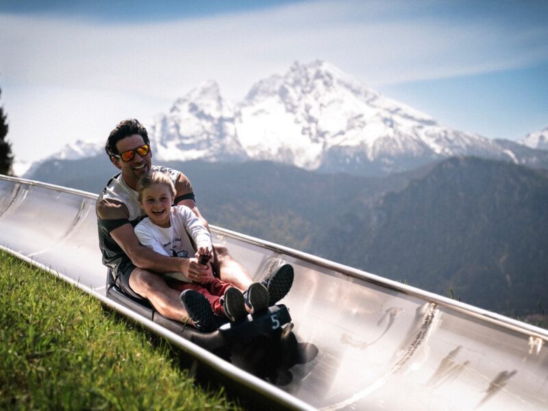 Vader en dochter lachend op de zomerrodelbaan bij Berchtesgaden met de besneeuwde Watzmann op de achtergrond, een spannend uitje met kinderen in Beieren