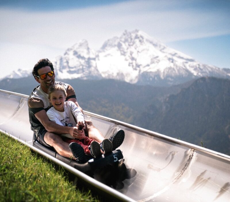 Vader en dochter lachend op de zomerrodelbaan bij Berchtesgaden met de besneeuwde Watzmann op de achtergrond, een spannend uitje met kinderen in Beieren