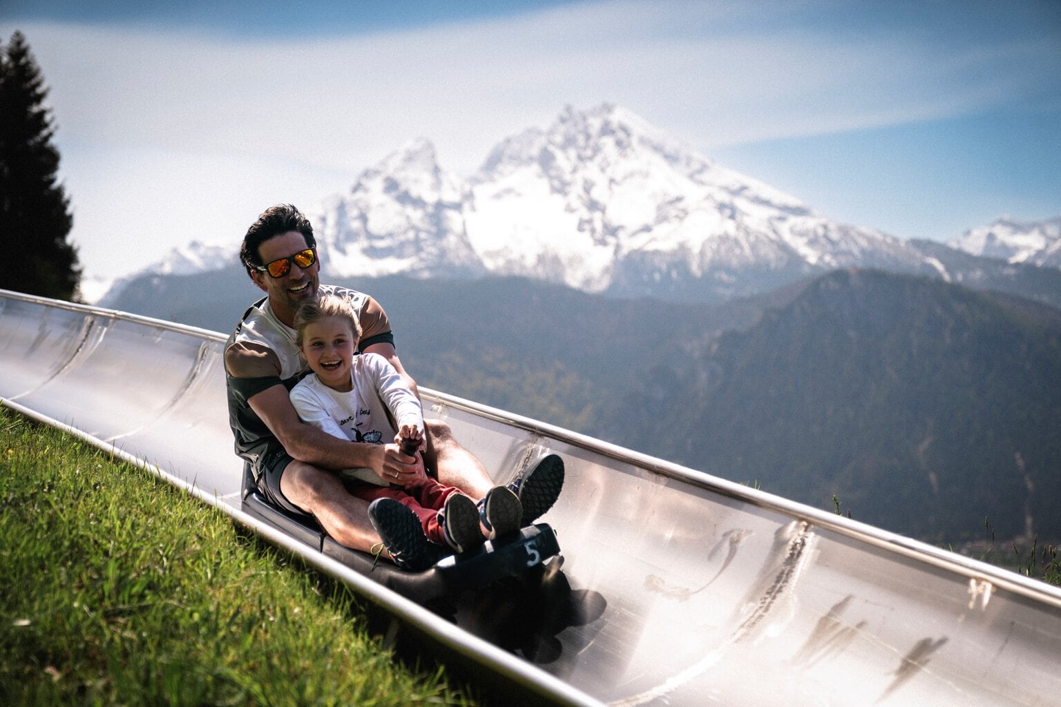 Vader en dochter lachend op de zomerrodelbaan bij Berchtesgaden met de besneeuwde Watzmann op de achtergrond, een spannend uitje met kinderen in Beieren